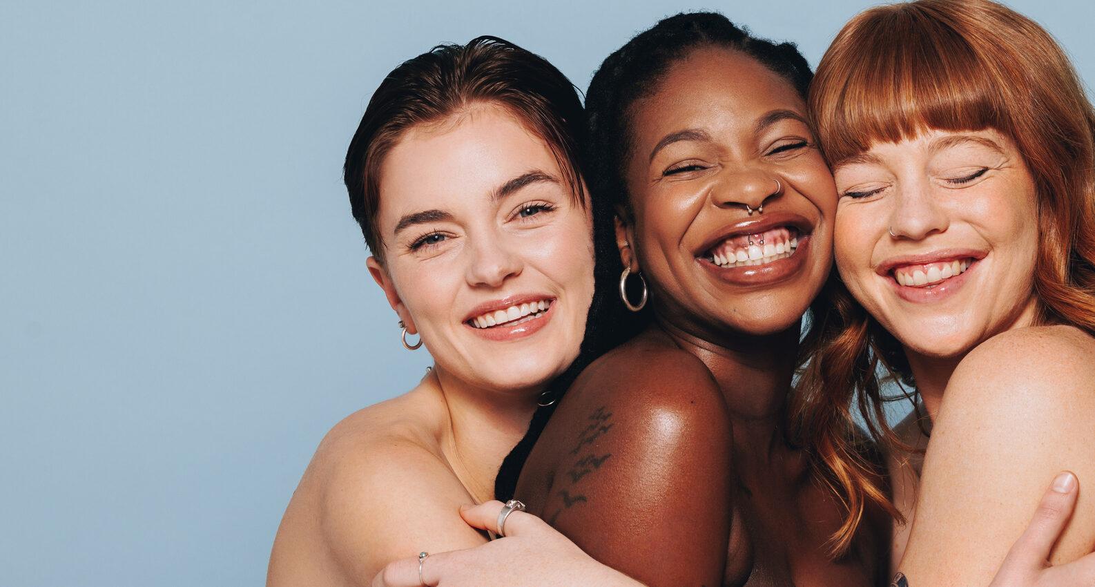 Group of happy women with different skin tones smiling and embracing each other. Three diverse women feeling comfortable in their natural skin. Body positive young women standing together in a studio.